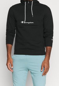 Felpa nera con zip e logo bianco "Champion" sul petto, dotata di cappuccio con cordino e polsini a coste. Abbinata a pantaloni blu chiaro.
