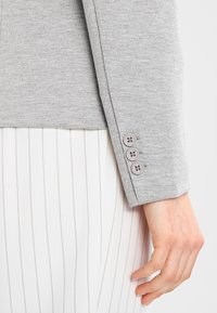 Šedý blazer s texturovaným povrchem a třemi knoflíkovými detaily na rukávu, kombinovaný s bílým pruhovaným oděvem pod ním.