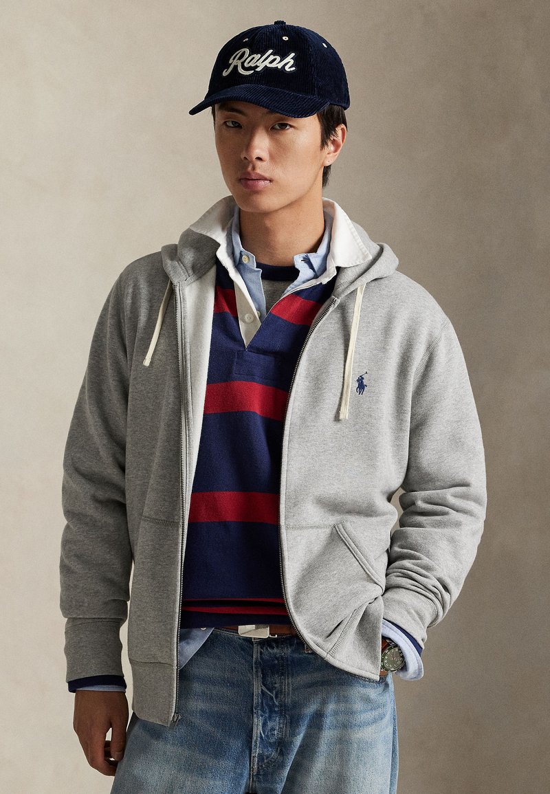 Junger Mann mit navyblauer Mütze, mehrlagigem Poloshirt mit roten und navyfarbenen Streifen, grauem Zip-Hoodie, hellblauem Hemd und blauen Jeans.