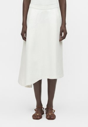 Žena nosi asymetrickú bielu midi sukňu v kombinácii s hnedými plochými sandálmi so remienkami, stojaca na jednoduchom bielom pozadí.