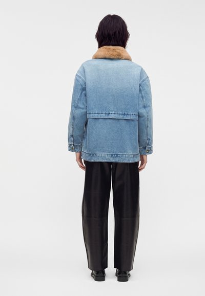 Personne debout de dos portant une veste en jean bleu clair avec un col en fourrure beige et un pantalon noir à jambes larges.