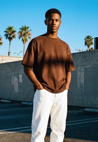 Fiatal férfi rövid hajjal, barna pólót és fehér nadrágot viselve, zsebeibe dugott kézzel áll egy napos, pálmafákkal övezett szabadtéri parkolóban.