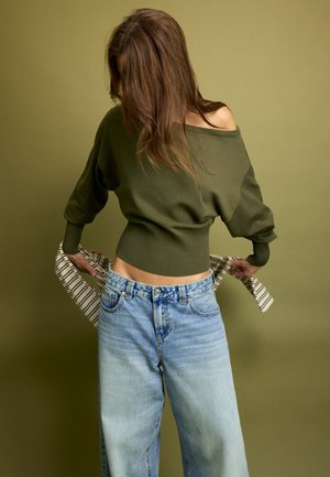Femme aux longs cheveux bruns portant un pull vert olive à épaules dénudées et un jean bleu clair, tenant une chemise rayée derrière son dos.