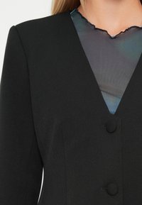 Zwarte op maat gemaakte blazer met stofbedekte knopen over een doorschijnende top met golvende randen in het donker, tegen een lichte achtergrond.