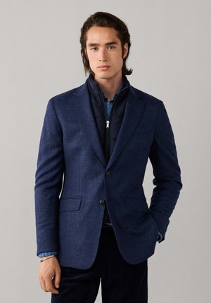 Joven con americana azul marino a cuadros sobre una chaqueta oscura con cremallera y un jersey azul, de pie con una mano en el bolsillo contra un fondo sencillo.