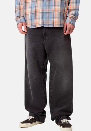 Persona con jeans neri oversize, sneakers nere con lacci bianchi e una camicia a quadri blu e arancione abbottonata.