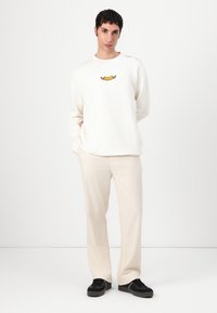 Krem genser med grafisk design i midten, kombinert med lysebeige bukser og svarte joggesko, plassert mot en nøytral bakgrunn.