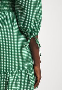 Zelené a bílé kostkované šaty s nafouknutými rukávy, lehce nařaseným trupem a zavázanými manžetami. Mají volánkový lem a lehkou bavlněnou látku.