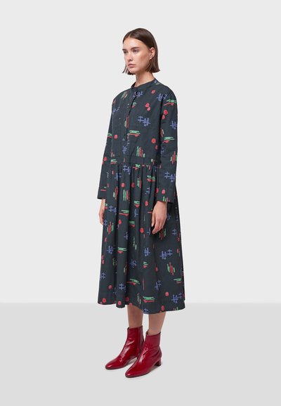 Vestido de manga larga hecho de tela oscura con un patrón geométrico multicolor. Tiene un escote redondo y una falda plisada. Se lleva con botas rojas hasta los tobillos.
