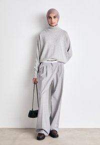 Hellgrauer oversized Pullover mit hohem Kragen, kombiniert mit hellgrauen, gefalteten Hosen. Das Model trägt eine schwarze Handtasche und braune Schuhe.