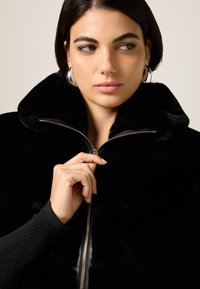 Mujer con cabello corto y oscuro cerrándose una chaqueta de piel negra, usando grandes aretes de argolla plateados, mirando ligeramente hacia la derecha.