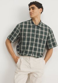 Chemise à manches courtes à carreaux dans des tons de vert foncé et blanc, avec un devant boutonné et deux poches poitrine, assortie à un pantalon couleur crème.