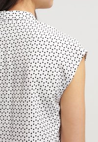 Kvinna iförd en ärmlös vit blus med ett svart oregelbundet rutmönster, sedd från baksidan och sidan mot en neutral bakgrund.