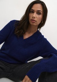 Pulover tricotat cu guler în V de culoare bleumarin, cu un finisaj texturat, mâneci lungi și manșete cu tiv, asortat cu blugi negri slim-fit.