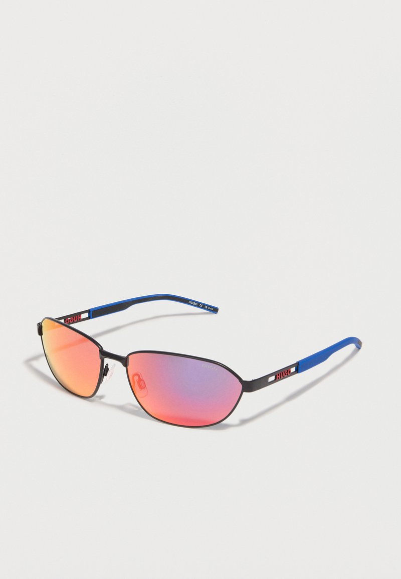 Γυαλιά ηλίου με μαύρο σκελετό, μπλε βραχίονες και ανακλαστικούς φακούς σε πορτοκαλί-ροζ, ακουμπισμένα σε φωτεινή επιφάνεια.