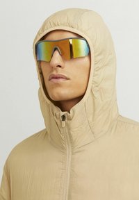 Beige hooded jacket with a zip closure, made from soft, textured fabric. Reflective, colorful sunglasses complete the look.