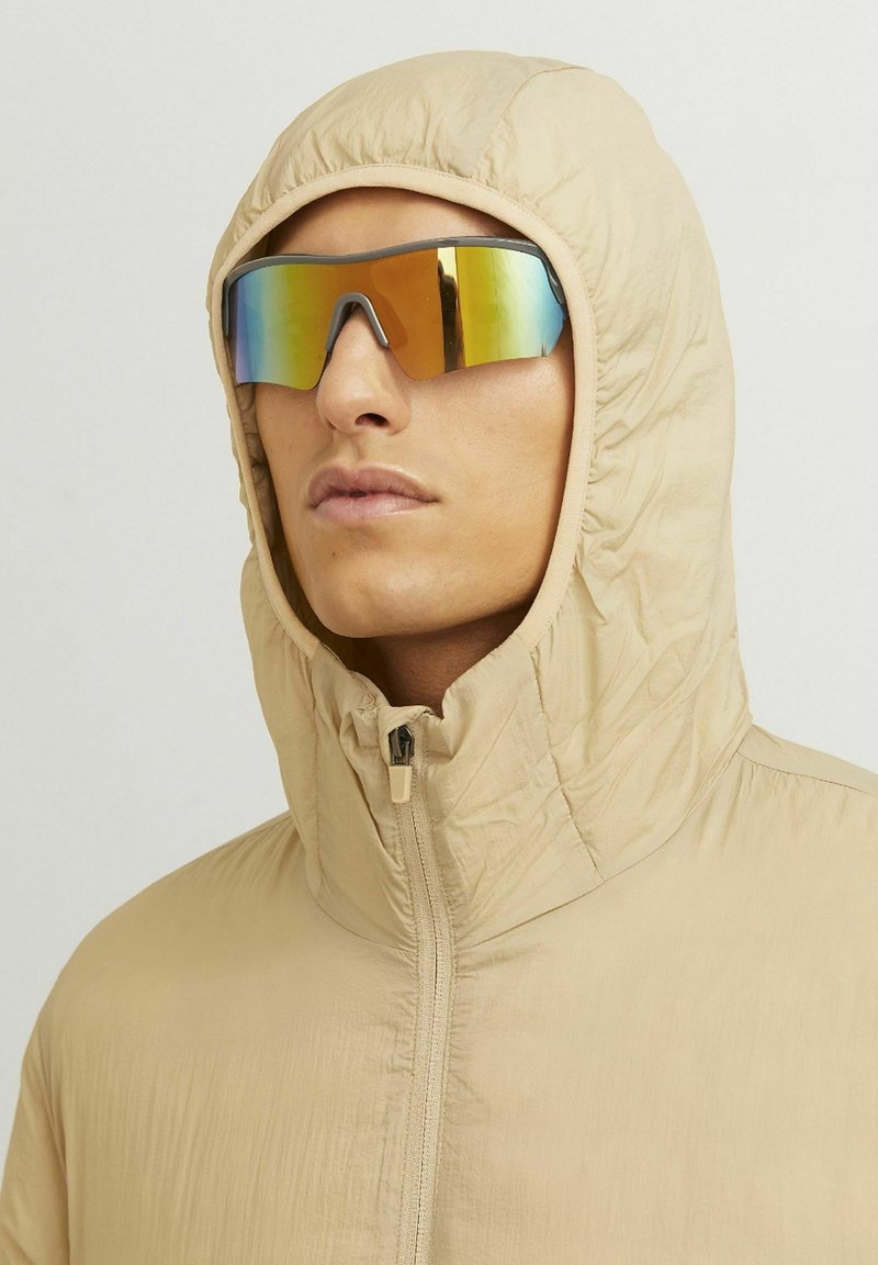Beige hooded jacket with a zip closure, made from soft, textured fabric. Reflective, colorful sunglasses complete the look.