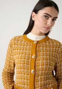 Mosterdgele cardigan met witte patroon, gestructureerde stof, ronde halslijn en vier metalen knopen aan de voorkant. Beschikt over voorzakken.
