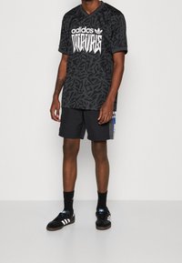 Camiseta negra de Adidas con patrón abstracto, logo blanco y escote en V. Combinada con pantalones cortos negros y zapatos negros con rayas blancas.