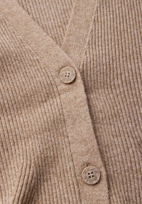 Cardigan beige lavorato a maglia con scollo a V, caratterizzato da coste verticali e chiusura a bottoni con quattro fori. Texture morbida con leggere variazioni di colore.
