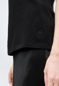 Κοντινό πλάνο ενός ατόμου που φοράει μαύρο πουκάμισο με ένα μικρό κεντημένο λογότυπο "S" κοντά στο τελείωμα και μαύρο παντελόνι, εμφανίζοντας το δεξί του χέρι.