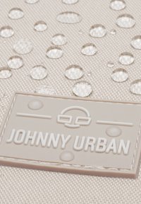 Water-repellent beige fabric with multiple water droplets, featuring a textured surface. Rubberized "JOHNNY URBAN" tag in a light gray color.