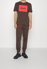 Hnědé bavlněné tričko s červeným potiskem ve tvaru krabice s nápisem "HUGO", doplněné o odpovídající hnědé tepláky a černé tenisky. Lehký, neformální outfit.