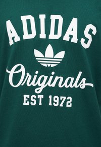 Πράσινο ύφασμα με λευκό λογότυπο Adidas, το κείμενο «Originals» σε γραφή και το «EST 1972» τυπωμένο από κάτω.