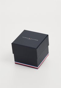 Tamno plava kartonska kutija s poklopcem, sa logotipom "TOMMY HILFIGER SATOVI" na vrhu i crvenom, bijelom i plavom trakastom dekoracijom na dnu.