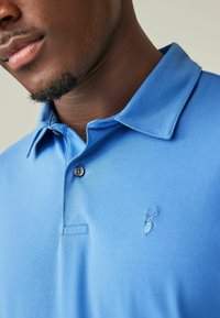 Helles blaues Poloshirt aus glattem Stoff mit einer Knopfleiste mit zwei Knöpfen, gesticktem Logo auf der Brust und einem klassischen Kragendesign.
