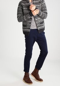 Cardigan de punto con patrón gris y negro sobre una camisa gris, combinado con pantalones azul marino y botas marrones con cordones.