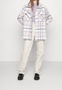 Geruite oversized shirt in lichtpaars, wit en zwart; twee borstzakken; gelaagd over een lavendeltop; gecombineerd met crèmekleurige broek en zwarte schoenen.