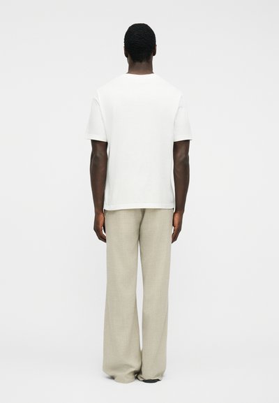 Άνδρας με την πλάτη, φορώντας απλό λευκό κοντομάνικο μπλουζάκι και φαρδιά μπεζ παντελόνια, μπροστά σε λευκό φόντο.