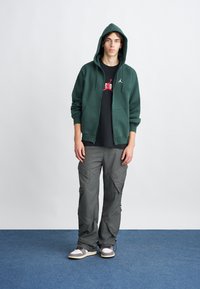 Felpa verde con zip e cappuccio regolabile, T-shirt nera con grafica rossa, pantaloni cargo grigi e sneakers grigie/bianche.