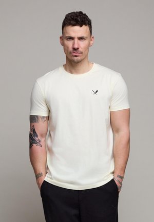 Mann med kort, mørkt hår iført en enkel off-white t-skjorte med en liten logo av kryssede cricketkøller og ball, svarte bukser, og synlige tatoveringer på armene.