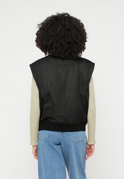 Colete preto sem mangas com uma textura lisa e brilhante e uma bainha canelada, usado sobre uma camisa de manga longa de cor verde-claro. Jeans de denim casuais completam o visual.