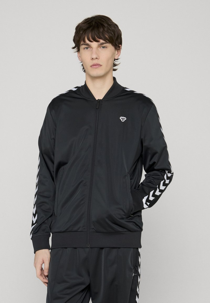 Veste de survêtement noire zippée en polyester lisse. Présente des bandes chevron blanches le long des manches et un petit logo sur la poitrine.
