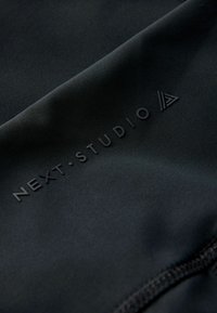 Crni sportski materijal s glatkom teksturom, s utisnutim logotipom "NEXT•STUDIO" i geometrijskim detaljima.