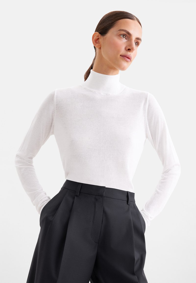 Femme aux cheveux foncés attachés en arrière, portant un pull à col roulé blanc et un pantalon noir large taille haute, les mains dans les poches.