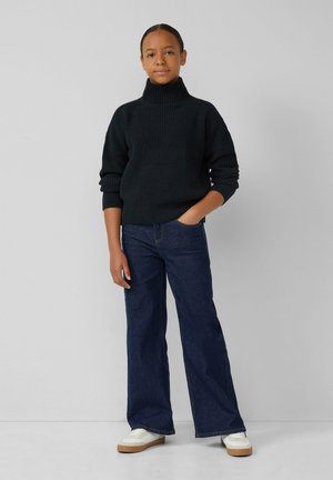 Donkergroene, geribbelde coltrui gecombineerd met wijde blauwe jeans en witte sneakers met beige zolen, handen in de zakken.