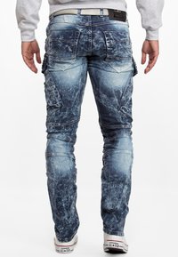 Pantalons cargo en denim bleu avec une finition usée, comportant plusieurs poches et des coutures visibles. Portés avec des baskets blanches.