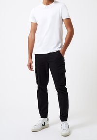 Camiseta blanca de cuello redondo, pantalones cargo negros con bolsillos laterales y zapatillas blancas con detalles verdes. Ajuste casual y textura suave.