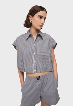 Mlada žena nosi košulju s kratkim rukavima, kopčanu, s okomitim prugama i kratke hlače s elastičnim pojasom u karo uzorku, stoji s rukama u džepovima.