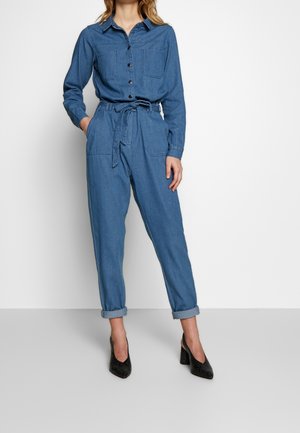 Frau trägt einen blauen Denim-Overall mit langen Ärmeln, Knöpfen vorne, Vordertaschen und Taillengürtel, kombiniert mit schwarzen Schuhen mit Absatz.
