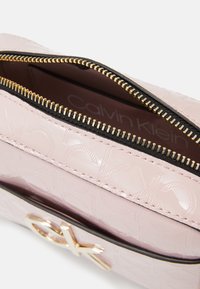 Jasnoróżowa saszetka Calvin Klein z wytłoczonym wzorem logo, częściowo otwarty złoty zamek błyskawiczny i złoty detal logo CK z przodu.