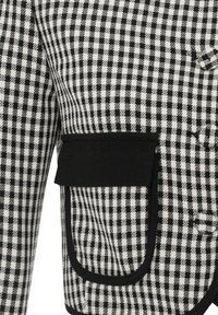 Veste à carreaux noir et blanc avec une grande poche bordée de noir et des boutons recouverts du même tissu sur le devant.