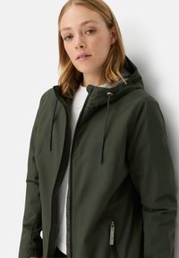 Olivengrøn vandtæt jakke med lynlås foran, justerbar hætte og snor, prydet med en subtil tekstur og sidelommer.
