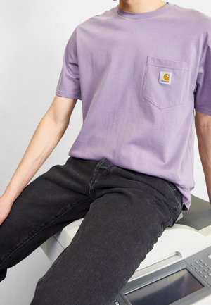 Person trägt ein lavendelfarbenes Carhartt-T-Shirt mit Tasche und schwarze Jeans, sitzt am Rand eines Kopierers oder Druckers.
