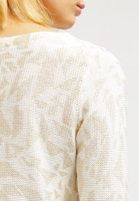 Une partie de l'épaule et du cou d'une personne portant un pull en tricot blanc avec un motif abstrait beige clair.