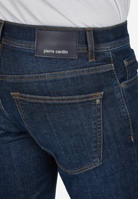 Dunkelblaue Jeans mit glatter Textur, mit einem Lederetikett mit dem Text "pierre cardin" und kontrastierenden orangen Nähten.